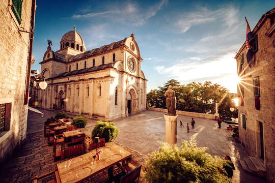 View of the Cathedral of St. James casting a shadow on the square with tables and chairs, statues, and people enjoying the sunny day, Sibenik, Sibenik-Knin County, Croatia.