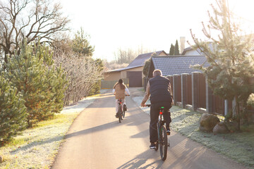 Fototapeta premium A man and teen cycle away down a sunlit rural path, frosty grass, evergreens, and long shadows evoke quiet companionship, seasonal transition, and shared peaceful journey.