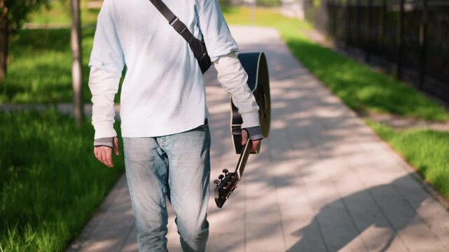 guitarist walking park path carrying guitar, young musician in white long sleeve shirt and jeans strolls along sunlit sidewalk holding black acoustic guitar by side with shoulder strap, sunlight