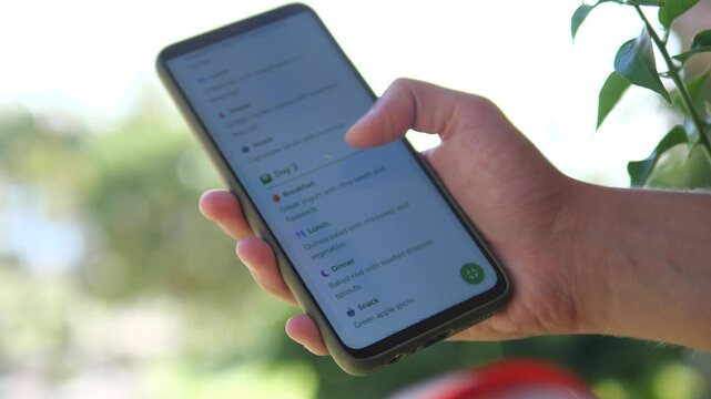 Close-up of hands scrolling through a personalized diet plan on a mobile screen. Tracking daily nutrition and healthy recipes in a bright garden. Modern technology for wellness, fitness goals, and