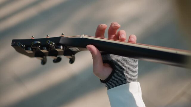left hand on guitar neck, closeup of lefthanded guitarist wearing grey knitted glove, sunlight and shadow patterns across fretboard, slowmotion fretting and tuning peg detail in quiet urban