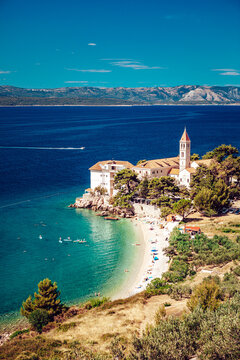 View of pristine turquoise waters gently kiss the shore of a white sandy beach nestled beneath an ancient stone church, Bol, Split-Dalmatia County, Croatia.