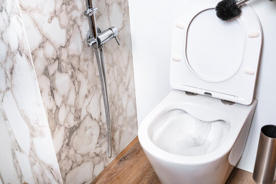 White ceramic toilet bowl with flowing water in stylish bathroom. Marble tile wall and bidet shower background. Minimalist interior design for home maintenance and hygiene blog
