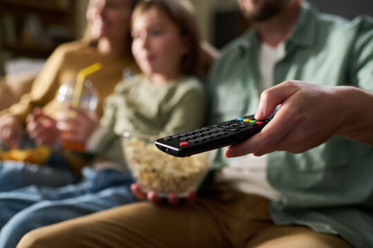 Caucasian man holding remote control while sitting with Caucasian woman and child on couch, watching television together, child holding drink and popcorn bowl visible