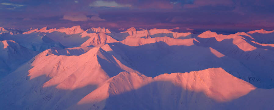 Aerial view of snow-capped peaks ablaze with the fiery hues of the setting sun, casting long, dramatic shadows across the pristine white landscape, Coldfoot, Alaska, United States.