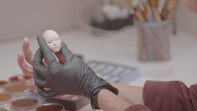 Artist holding silicone doll face with hands while inspecting fine features, surrounded by paints and tools, showing focus on miniature detailing work