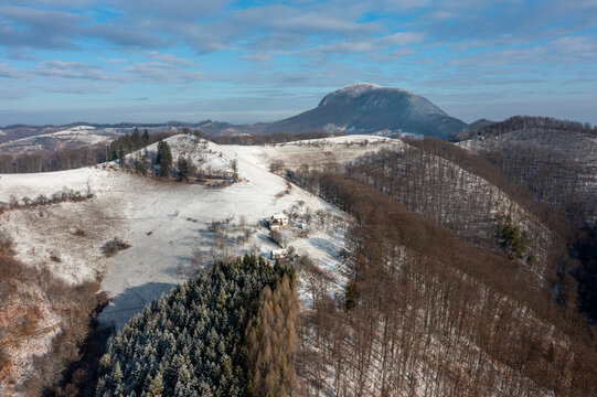 Aerial view of snow-dusted hills and forests create a stark contrast under a bright sky, with a majestic mountain in the background, Holbav, Brasov, Romania.