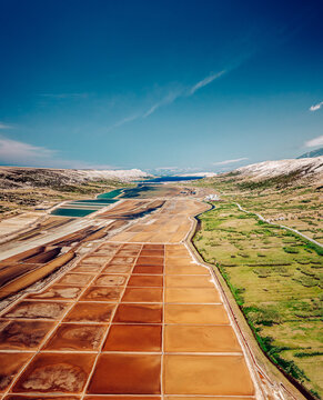 Aerial view of vibrant salt pans painted in earthy tones contrasting with the lush green fields under a clear blue sky, Pag, Zadar County, Croatia.
