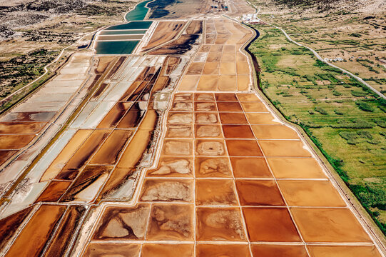 Aerial view of patterned salt pans reflecting the sky, contrasting with the natural green landscape, Pag, Zadar County, Croatia.