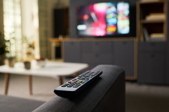 Remote control resting on sofa armrest with blurred television screen in background showing colorful content, modern living room setting with coffee table and shelves visible