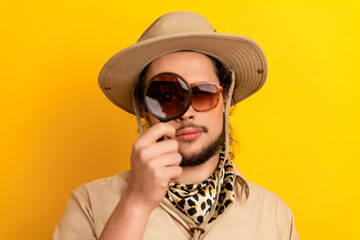 Obraz premium Man with safari hat and leopard scarf examines scene with a magnifying glass against a bright yellow background