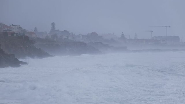 Large waves crash against a rocky shoreline during foggy stormy weather, with coastal houses and cliffs in the distance.