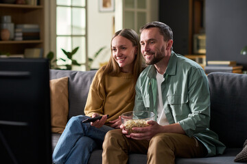 Fototapeta premium Caucasian couple sitting on sofa watching television together, smiling and holding bowl of popcorn, enjoying leisure time at home