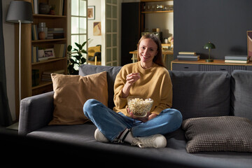 Obraz premium Portrait of young adult woman sitting cross legged on sofa eating popcorn and smiling while watching television at home, holding bowl in lap, relaxed posture, casual setting
