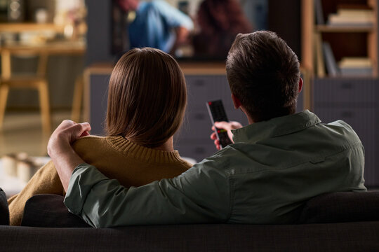 Caucasian young adult man and Caucasian young adult woman sitting on sofa watching television together, man holding remote control, couple relaxing at home, backs facing camera