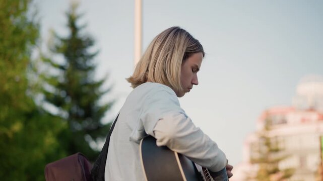 blonde white guitarist tuning acoustic guitar outdoors in city plaza, closeup hands and fingers adjusting capo and strings, backpack and strap visible, focused rehearsal and strum practice, indie