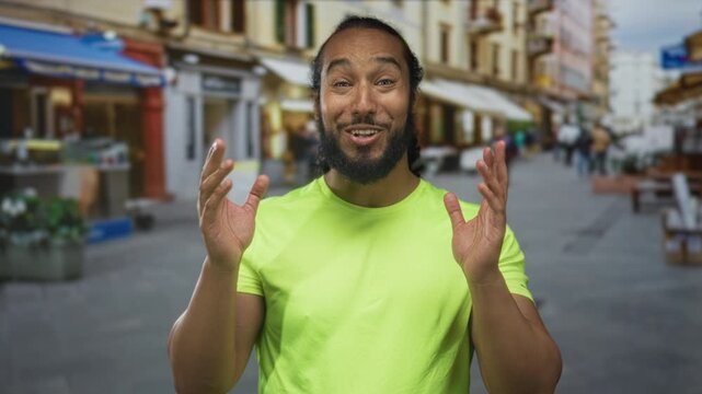 Man with hand on chest standing on a busy street, smiling toward camera in neon green shirt with beard and braided hair; gratitude connection.