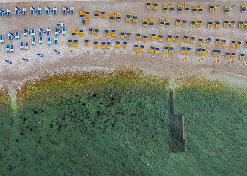 Aerial view of orderly rows of beach umbrellas casting shadows on the sandy shore where the sea transitions from clear turquoise to deep blues, Portonovo, Provincia di Ancona, Italy.