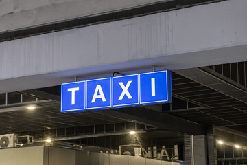 Bright blue illuminated taxi sign with white letters hanging from an urban structure, indicating transportation services and convenient traveler pick-up guidance
