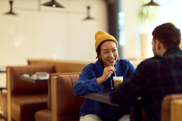 Asian Woman in Yellow Beanie Enjoying Coffee Chat With Friend in Cozy Cafe Sitting Together