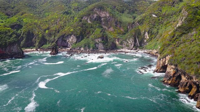 Aerial drone view of a grass-covered sea stack with a sea cave on Chilo&eacute; Island, Patagonia&rsquo;s Pacific coast, Chile. Powerful waves hit a remote sandy beach and rugged green cliffs under an overcast sky