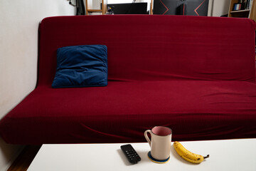 Red couch with an blue velvet pillow, low table in front holding a coffee cup, remote, and banana. Casual, everyday living room scene with copy space. Concept of home, comfort, relaxation, lifestyle