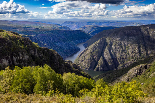 Aerial view of the serpentine river snaking through the deep, shadowed gorge, flanked by verdant hills under a sky brushed with clouds, Canyon du Sil, Lugo, Spain.