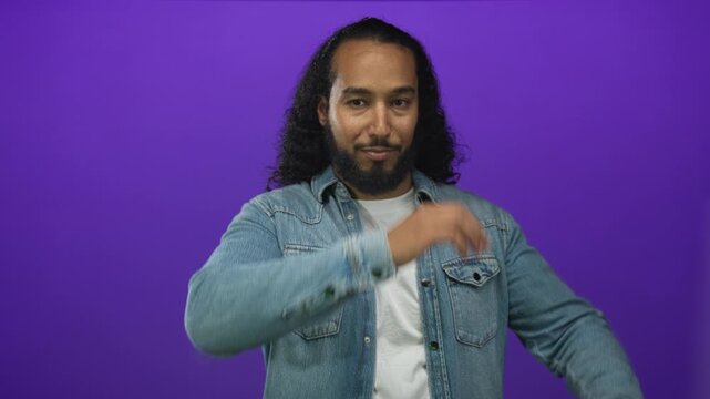 Man presenting with hand and open palm in purple studio wearing denim jacket and white tee with direct gaze toward camera; confidence focus.