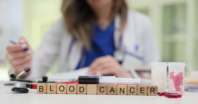 Wooden cubes spell Blood Cancer near syringe and bright red pills. Female doctor writes report about blood cancer diagnosis on desk in clinic