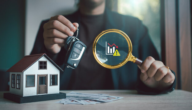Businessman hand holding car key and magnifying glass with virtual risk warning graph icon over house model and money for financial crisis management concept, Economic recession Inflation Repossession