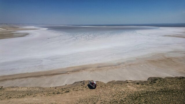 Aerial view of the vast, sun-drenched salt flats stretching to the horizon under a clear blue sky, meeting the rugged coastline, Say-Utes, Mangystau Region, Kazakhstan.