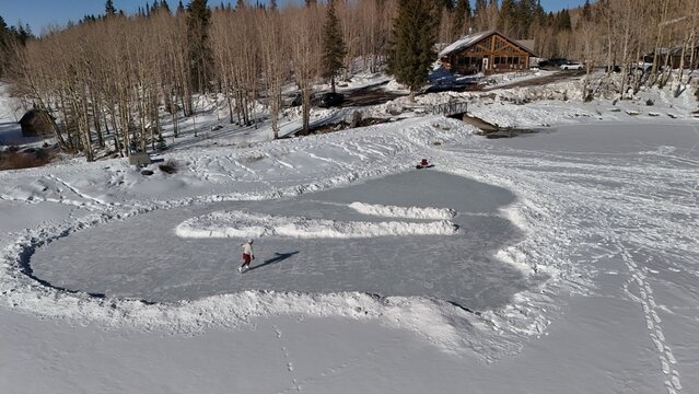 Aerial view of skaters gliding across a frozen expanse, framed by snow-dusted trees and a cozy cabin nestled along the shore, Mesa, Colorado, United States.