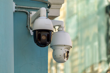 Two dome security cameras mounted on exterior wall with visible conduit and wiring. Surveillance equipment points in different directions near building corner.