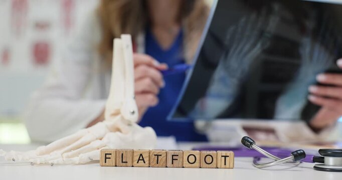 Wooden cubes spell Flatfoot standing near bone model of ankle and toes on table. Female doctor studies foot X-ray checking foot deformity on image