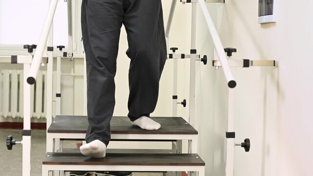 A man undergoes rehabilitation after an injury on a stair climber with parallel bars at a clinic under the supervision of a physical therapist. Close up legs on stairs
