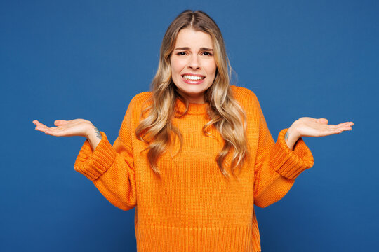Young sad Caucasian woman she wears yellow sweater casual clothes shrugging shoulders looking puzzled, have no idea spread hands isolated on plain blue background studio portrait. Lifestyle concept.