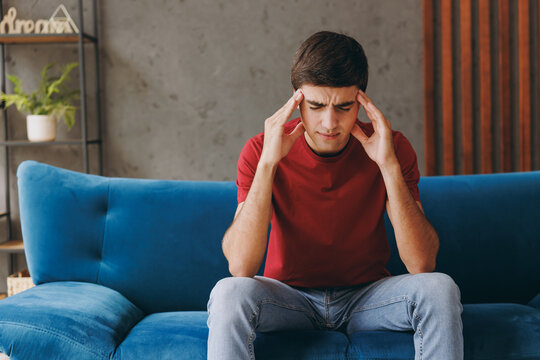 Young pensive dissatisfied depressed sad man wear red t-shirt hold temples with hands sit on blue sofa couch stay at home hotel flat spend free spare time in living room indoors People lounge concept