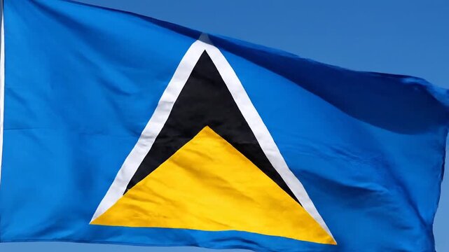 The national flag of Saint Lucia, featuring a blue field and a central triangular design (yellow, black, and white), waves proudly and vigorously in the wind against a clear, sunny blue sky.