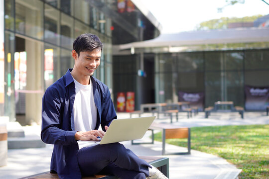 Handsome asian business man or student typing on laptop sitting on bench in modern urban environment
