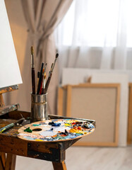 Artist's workspace canvas on easel, palette with paint, brushes, and natural light from a window