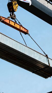 Vertical video: Hoist engaging, spreader lifting steel panel at construction site, flag fluttering