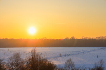 Obraz premium Picturesque winter landscape with a bright sun rising over a snowy field and a forest line. Warm golden light dissipates the morning mist, creating an atmosphere of peace and harmony in the wild natur