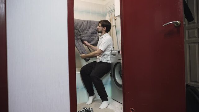 Caucasian man beating down jacket on drying rack in bathroom to evenly distribute down after washing. Life hack for smoothing filler inside down jacket.