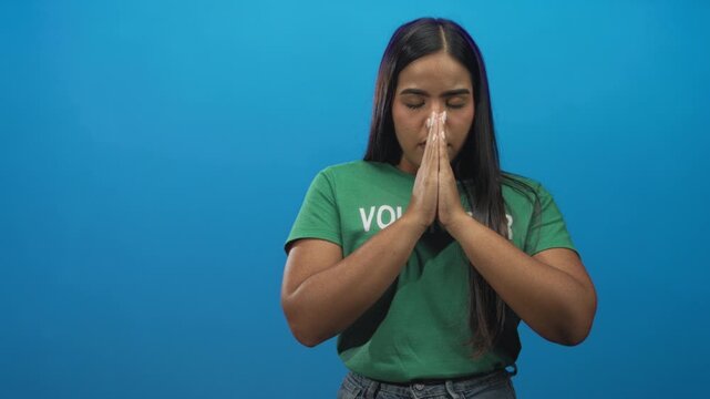 Young hispanic volunteer woman with hands pressed together in prayer and green tshirt in studio; serene reflection solidarity.
