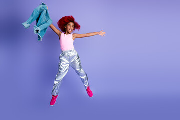 Young girl in a pink top and metallic pants leaps with denim jacket in hand against a purple studio...