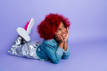 Happy young girl with red dyed hair lying on purple background wearing denim jacket and metallic...