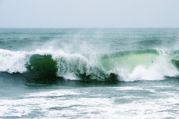 Obraz premium a single powerful green wave cresting and breaking with misty white spray against a grey sky.