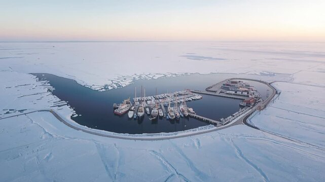 Gliding drone revealing marina with moored boats, breakwater and icy channel, documenting harbor