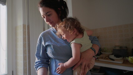 Mother holding curly-haired toddler on hip in cozy kitchen with soft window light, both gazing...