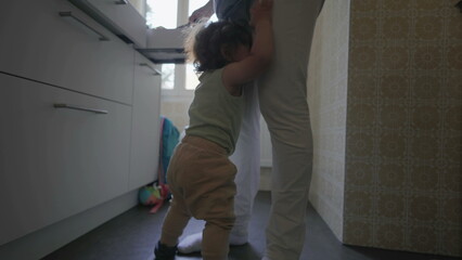 Toddler clinging to parent&rsquo;s leg in kitchen with arms raised and complaining expression, asking to be picked up while parent stands holding phone nearby